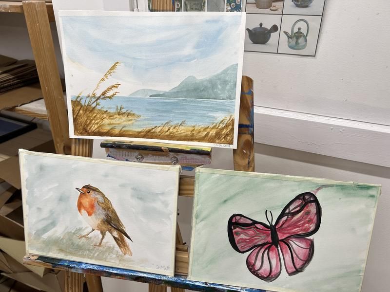 Student watercolours of a robin, a butterfly and a lakeside landscape on easels