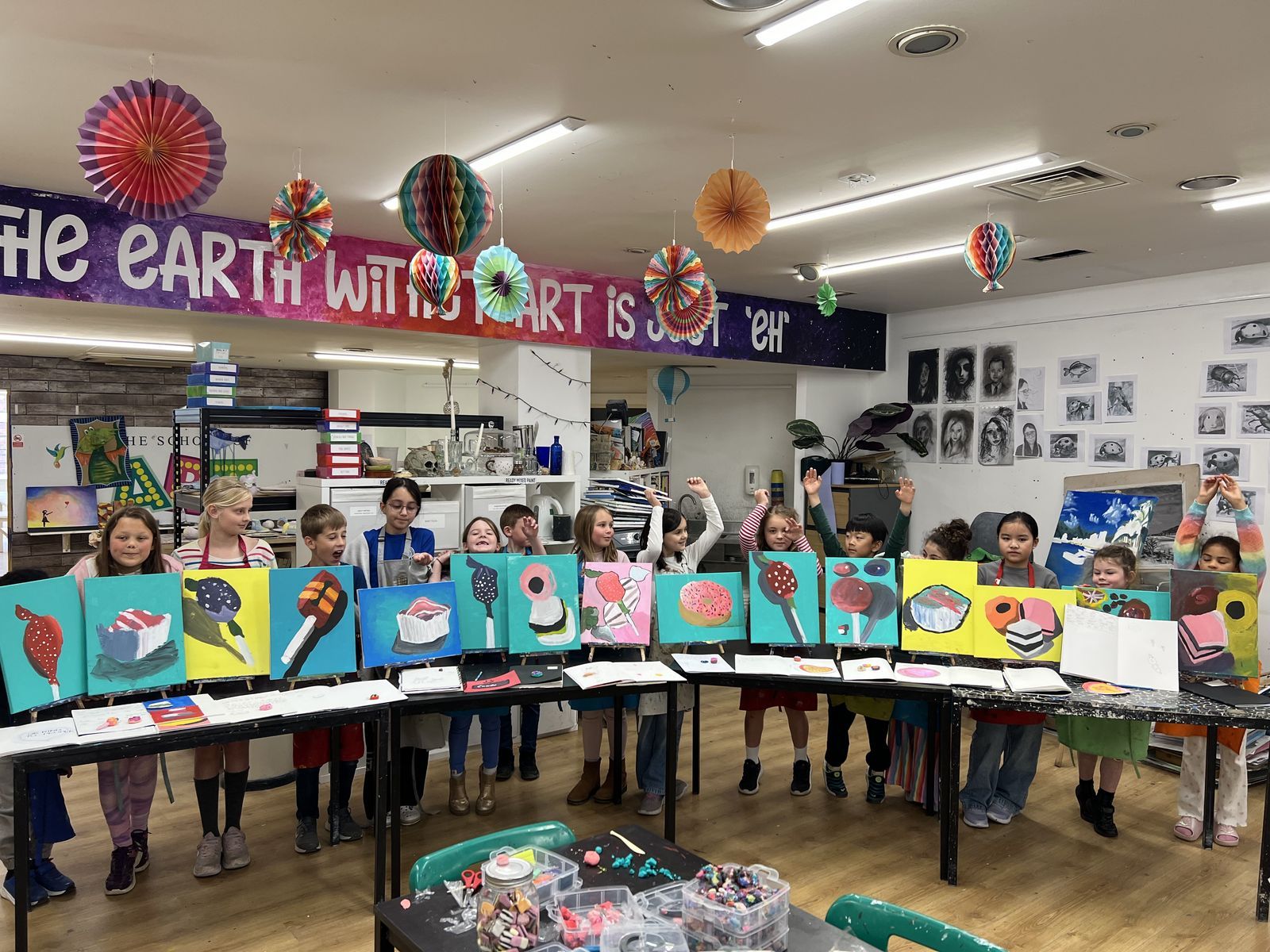 A full class of children in aprons cheering behind a long row of their finished colourful paintings in The Art School studio