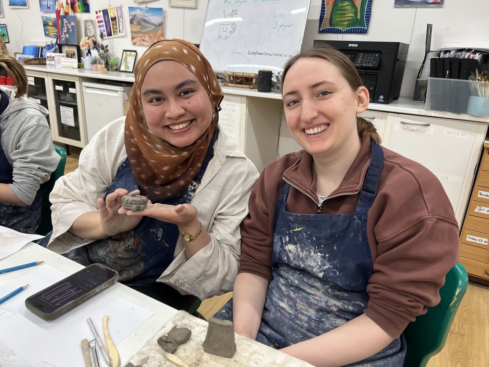 Two smiling students in aprons showing the small clay pieces they are making at a pottery workshop